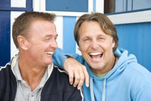 A colour portrait photo of a happy laughing gay male couple having fun together while sitting in front of some beach huts.